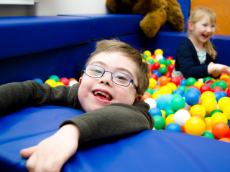 Ein kleiner Junge mit Downsyndrom hält sich lachend am Rand eines Bällebades fest