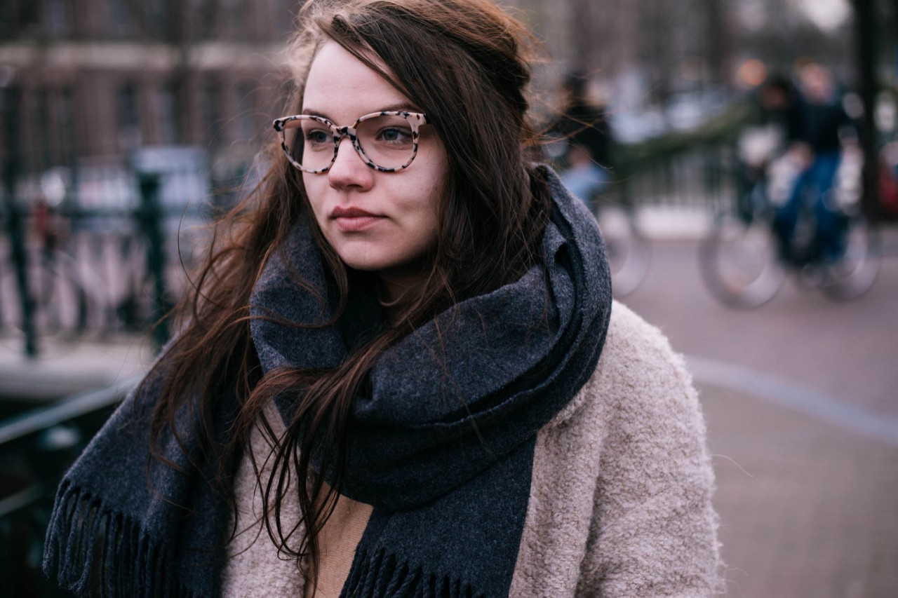 Junge Frau mit dickem Schal und Brille in st&auml;dtischer Umgebung schaut nachdenklich