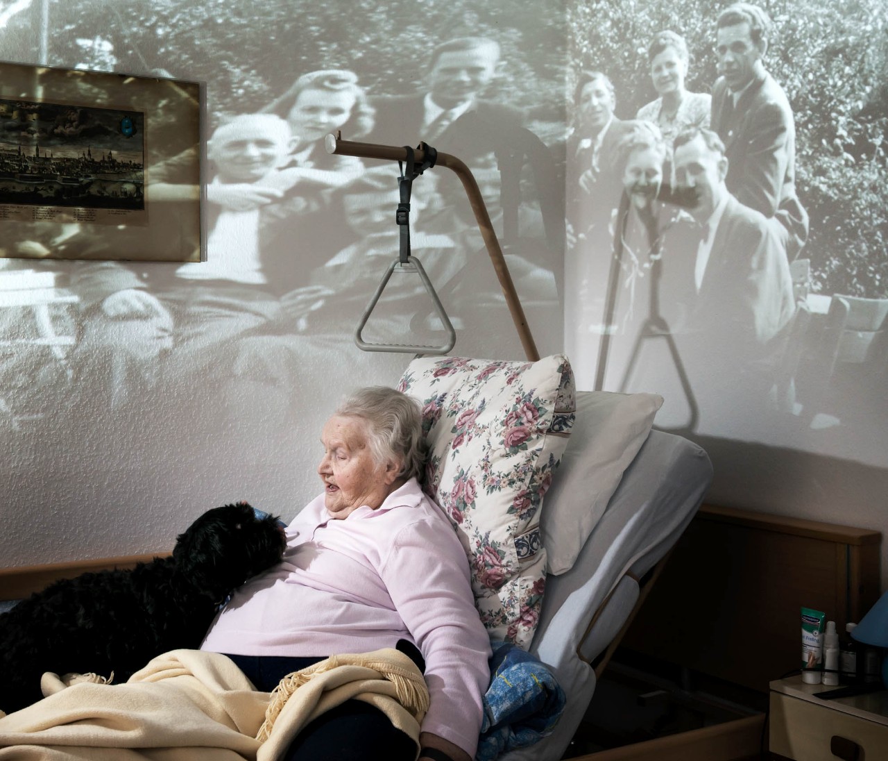 Eine alte Frau liegt in einem Pflegebett. An die Wand hinter ihr ist ein altes Foto projiziert.