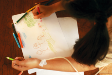 Junges Mädchen malt am Tisch ein Kinderbild, in ihrem Arm ein dünner Infusionsschlauch.