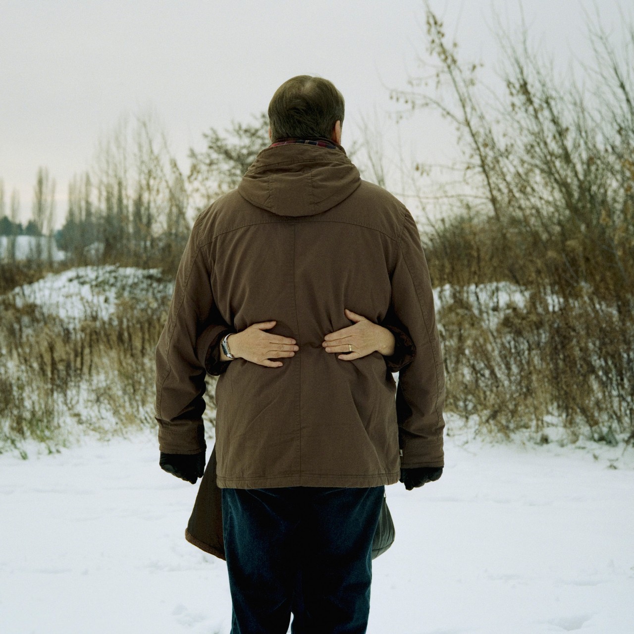 Ein Mann steht mit dem Rücken zur Kamera in Winterlandschaft. Auf dem Rücken zwei Hände von einer Frau, die ihn umarmt.