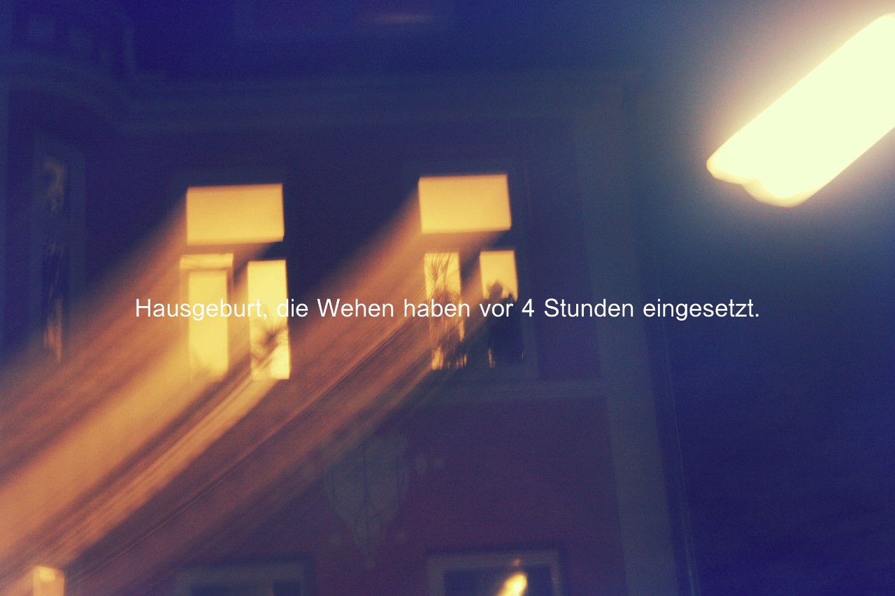 Zwei erleuchtete Hausfenster bei Nacht, davor die Aufschrift "Hausgeburt, die Wehen haben vor 4 Stunden eingesetzt."