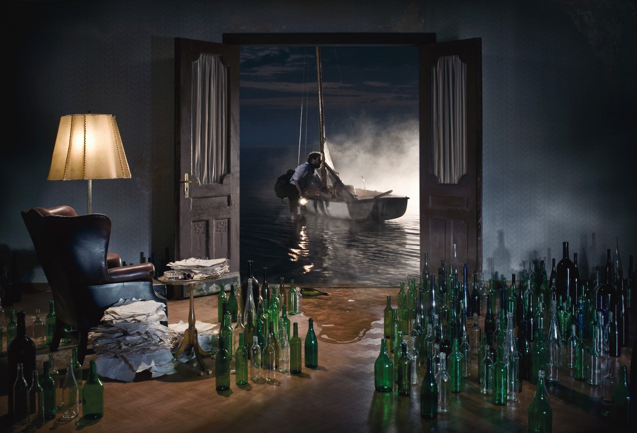 Surreale Fotomontage eines Wohnzimmers mit leeren Flaschen, hinter einer offenen Flügeltür Wasser mit einem Segelboot bei Nacht.