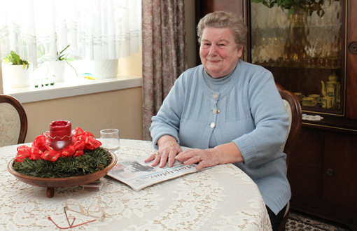 Ältere Frau sitzt an einem Wohnzimmertisch am Fenster und lächelt zufrieden