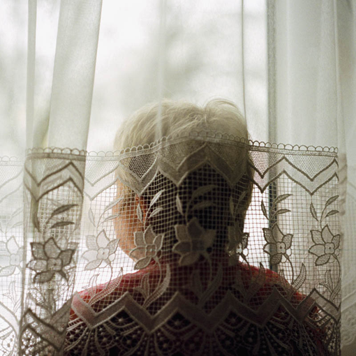Rückansicht einer älteren Frau, die sich zwischen die Fensterscheibe und die durchsichtigen Gardinen gestellt hat und auf eine Winterlandschaft blickt.