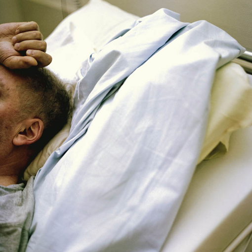 Ausschnitt vom Kopfende eines Krankenbettes, von einem älteren Mann ist nur der seitliche Kopf ohne Gesicht und eine Hand auf der Stirn sichtbar.