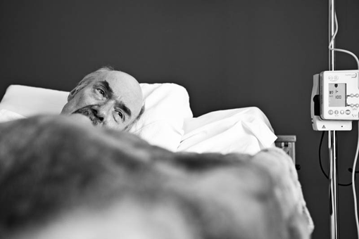 Blick auf ein Krankenbett, vom Kranken ist nur das Gesicht erkennbar, daneben ein Infusionsständer.