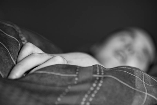 Portrait eines Jungen Manns im Bett. Im Vordergrund scharf die Hand auf der Bettdecke, im Hintergrund verschwommen das Gesicht.