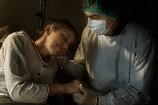 Eine Frau im Bett zur Seite aufgestützt, neben ihr hält ein Arzt mit Atem- und Haarschutz ihre Hand.