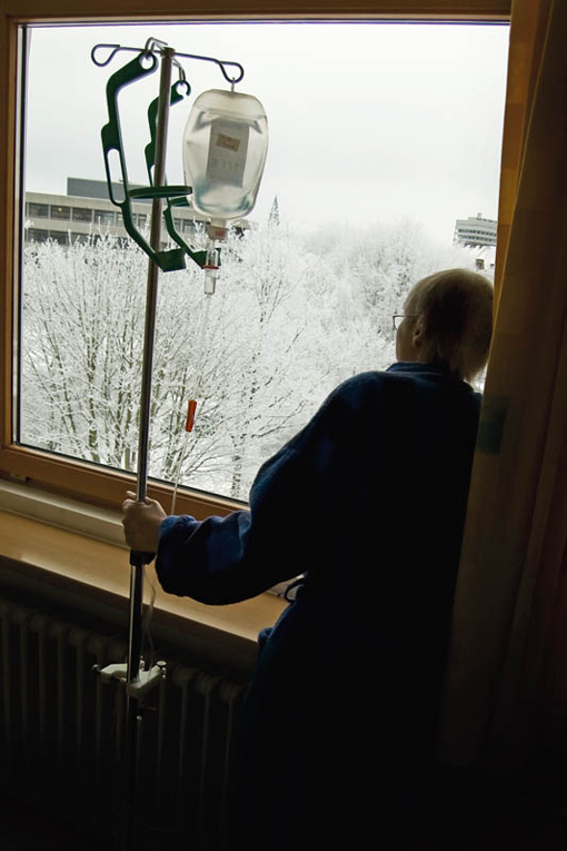 Eine Frau blickt aus dem Fenster in eine Schnee-Landschaft und hält neben sich ein mobiles Infusionsgerät.