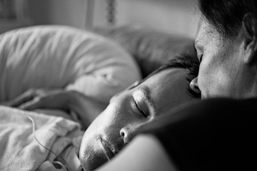 Portrait eines im Bett liegenden jungen Mannes, eine ältere Frau lehnt ihr Gesicht auf seine Stirn, beide mit geschlossenen Augen.